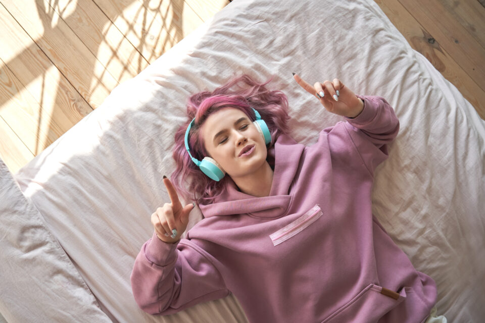 Happy funny teen girl with pink hair wear headphones lying in comfortable bed listening new pop music enjoying singing song with eyes closed relaxing in cozy bedroom at home. Top view from above. - Nők Lapja