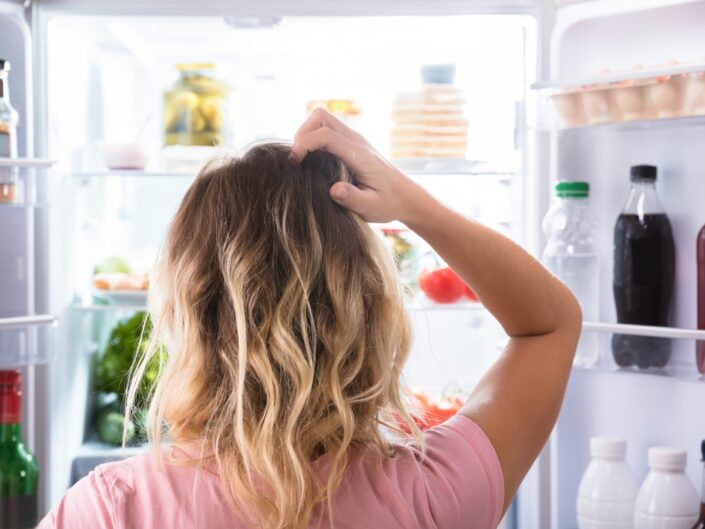 egy nő a nyitott hűtőszekrény előtt áll, és a fejét vakarja