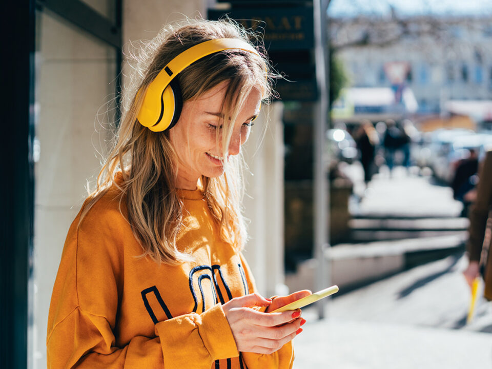Close-up portrait of smiling happy caucasian woman scrolling on social media and listening to audio with wireless headphones - Nők Lapja mobilzombik-okostelefon