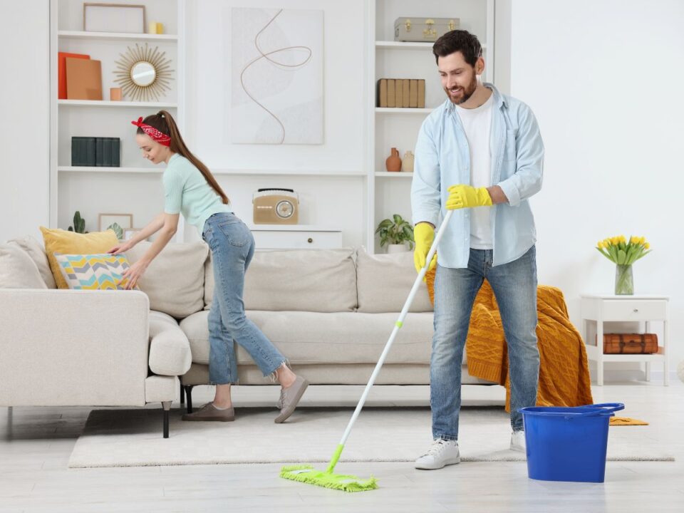 rendrakas-pszichologia-boldogsag - Nők Lapja világos nappaliban balra hátrébb egy nő rendezgeti a kanapé párnáit, jobbra elöl egy férfi épp felmossa a parkettát, mindketten jókedvűek