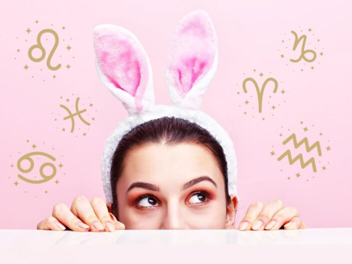 Egy nő rózsaszín háttér előtt nyuszi füles fejpántban kukucskál fel egy felület mögül, körülötte arany színű csillagjegy szimbólumok lebegnek.