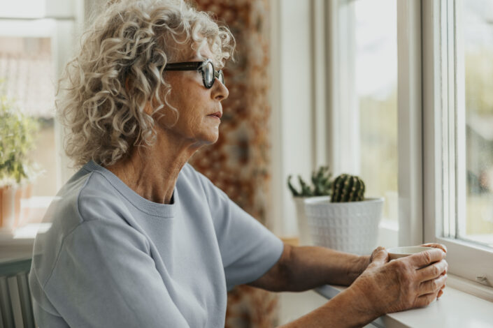 szőke középkorú szemüveges nő ablak előtt ül mereng kifelé néz