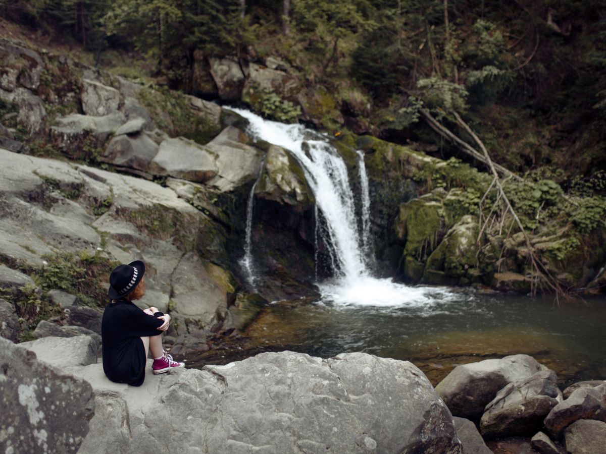 Portré egy fiatal kaukázusi nőről, aki egy vízesés előtt ül.