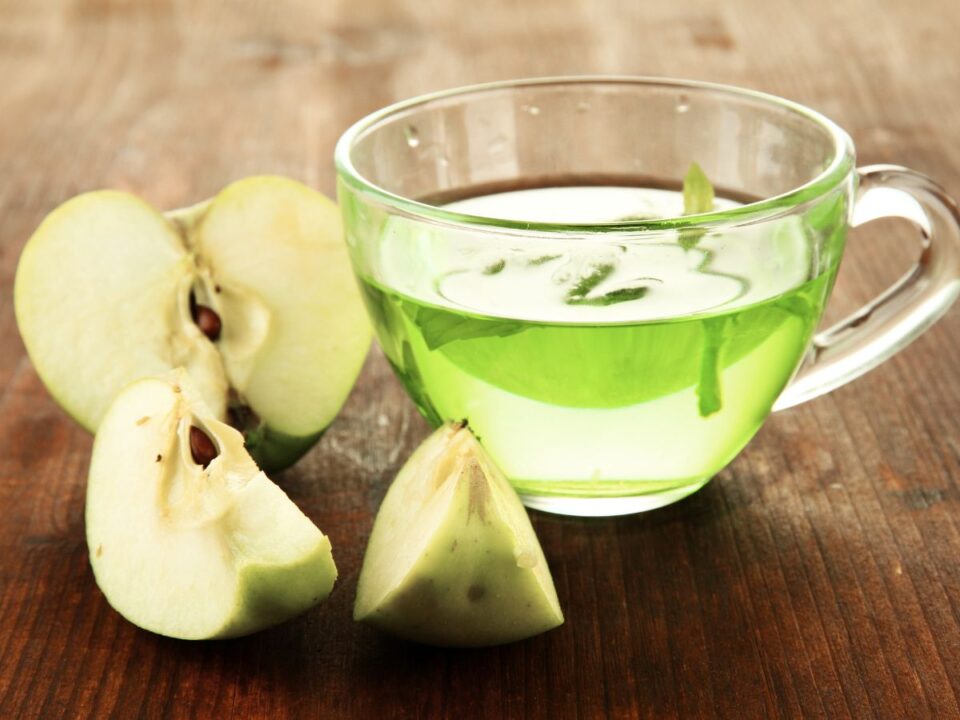 barna fa asztalon balra szeletelt zöld alma, jobbra átlátszó csészében zöld ital