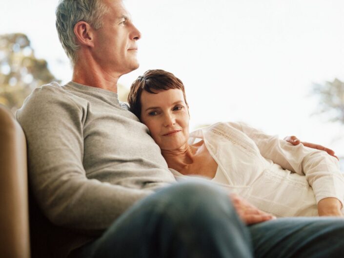 Ötvenes éveiben járó férfi és nő kanapén ül egymást átölelve, nő kinéz a kamerába