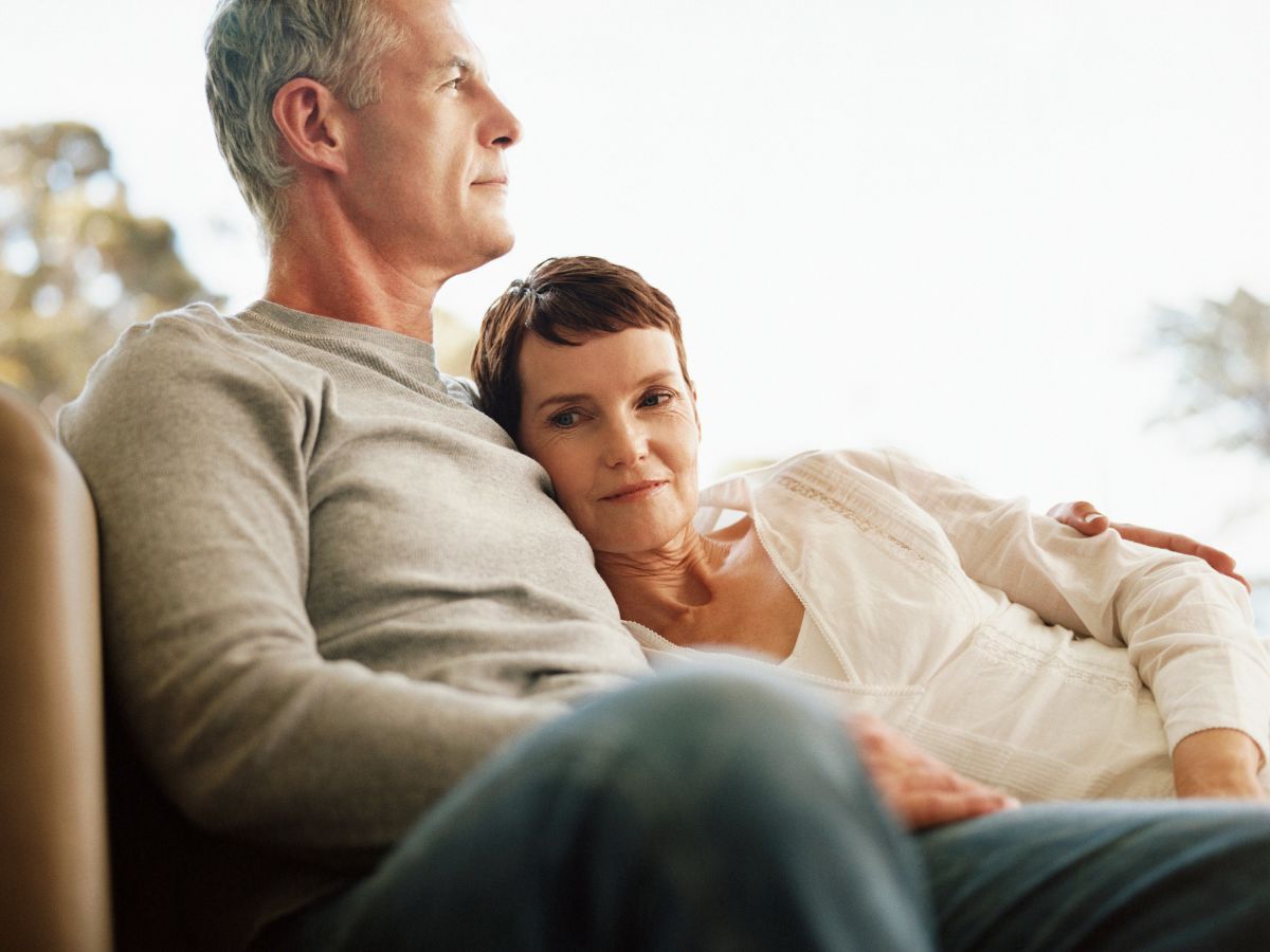 Ötvenes éveiben járó férfi és nő kanapén ül egymást átölelve, nő kinéz a kamerába
