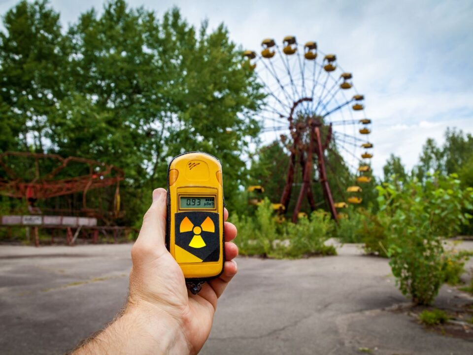 csernobili-gomba-sugarzas-titka - Nők Lapja Egy kéz sugárzásmérőt tart egy elhagyatott vidámparkban; háttérben rozsdaette óriáskerék és benőtt növényzet a csernobili zónában.