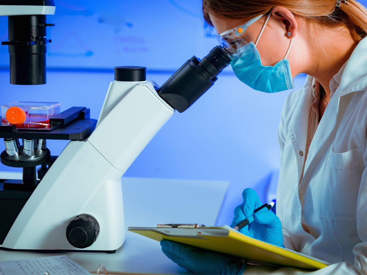 Egy védőfelszerelést viselő női kutató mikroszkópba néz laborban, jegyzetel közben; kék fényű környezet, steril eszközök és minták láthatók.