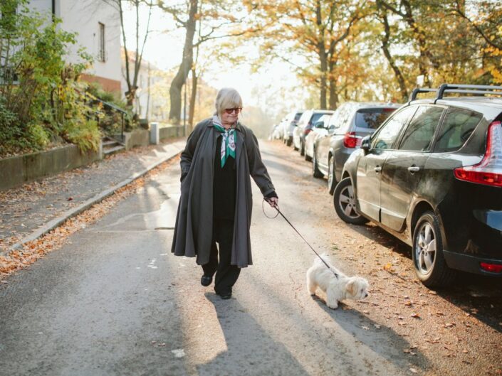 középkorú nő ballonkabátban reggel kutyát sétáltat az utcán