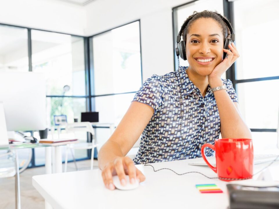 világos irodában egy nő fejhallgatóval a fején mosolyogva dolgozik a számítógépén, rajta csíkos póló, keze az egéren,az asztalán egy narancsárga bögre