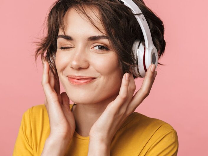 fiatal, barna hajú nő sárga pólóban, fehér fejhallgatóval a fülén kacsint, a háttér rózsaszín