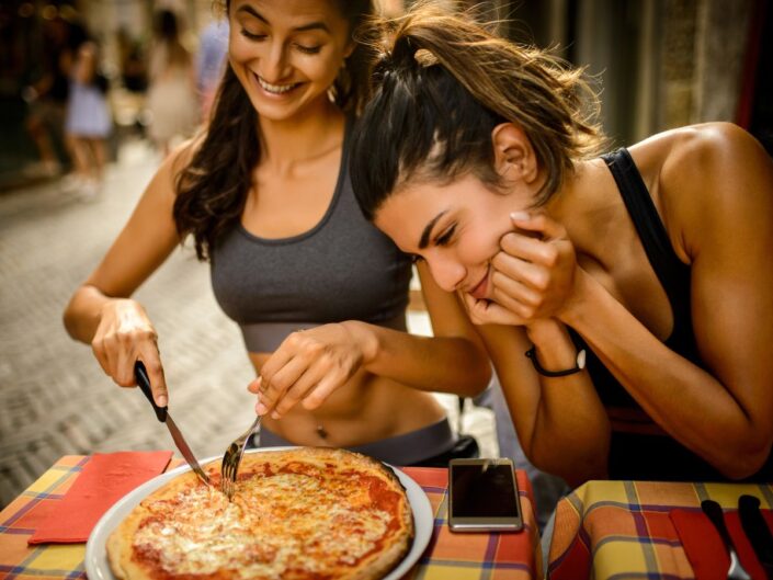 Sportos testalkatú fiatal nő, mosolyogva vágja fel maga előtt az asztalon lévő pizzáját, egy másik nő fölé hajol és vágyakozva nézi a pizzát