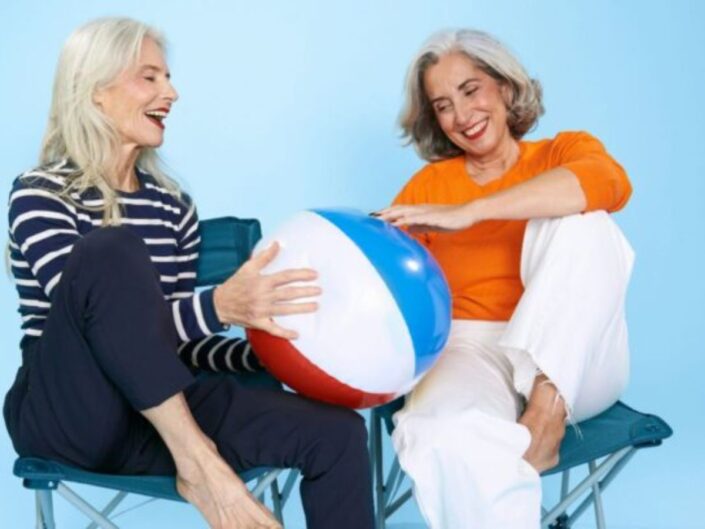 Egy hosszú, ősz hajú, szép idősödő nő tengerészcsíkos felsőben és kék nadrágban, és egy szép, bob hajú idősödő nő narancsszínű felsőben, fehér nadrágban kék háttér előtt horgásszékben vidáman strandlabdát fog