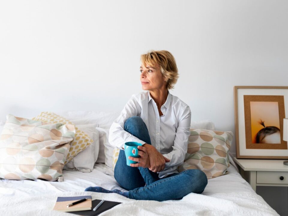 nő farmerban ül az ágyon és kávézik