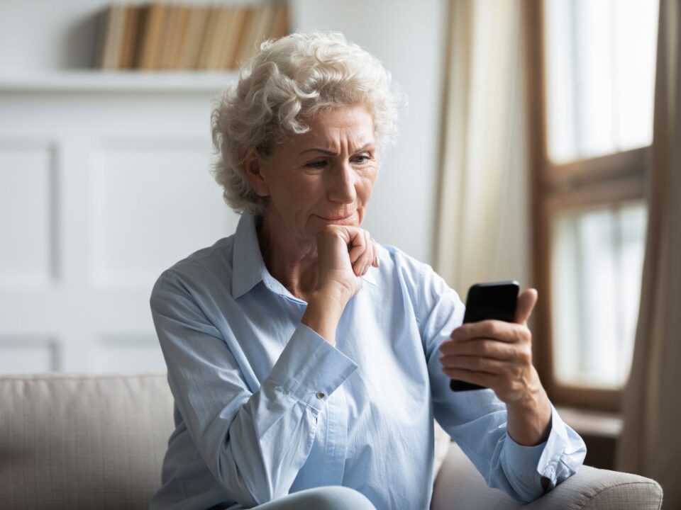 Egy idősebb nő fotelben ül, kezében okostelefont tart, miközben elgondolkodva nézi a képernyőt. Arckifejezése komoly, kissé aggódó, a háttér világos és csendes otthoni környezetet mutat.