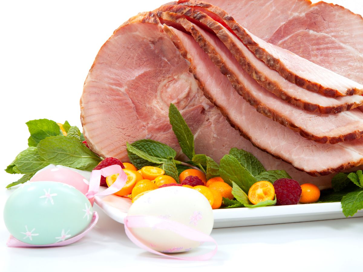 Szeletelt főtt sonka egy tálon, mellette festett húsvéti tojások, zöld levelek és apró gyümölcsök, ünnepi, világos háttérrel.