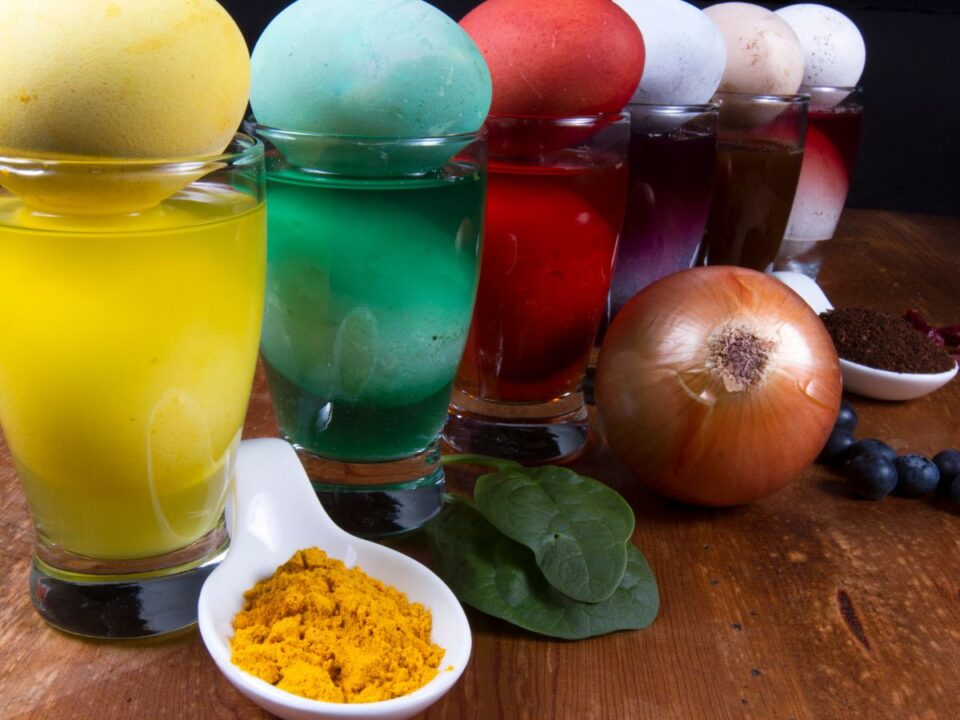 Poharakban különböző színű festőlevek sorakoznak, rajtuk tojások. Elöl kurkuma, hagyma és zöld levelek láthatók, bemutatva a természetes tojásfestés alapanyagait.