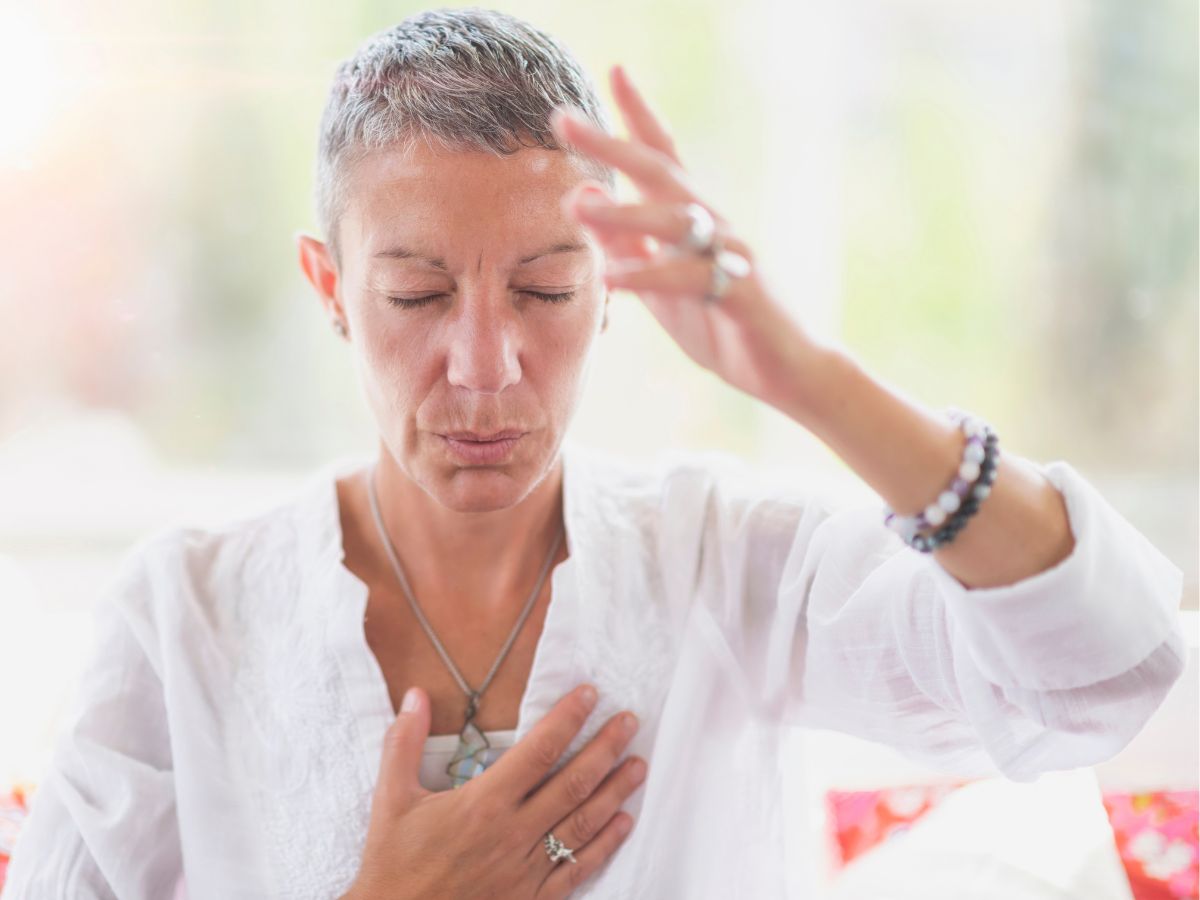 fehér blúzos, rövid hajú, idős nő jobb kezével a mellkasát, bal keze hüvelykujjávala homlokátérinti