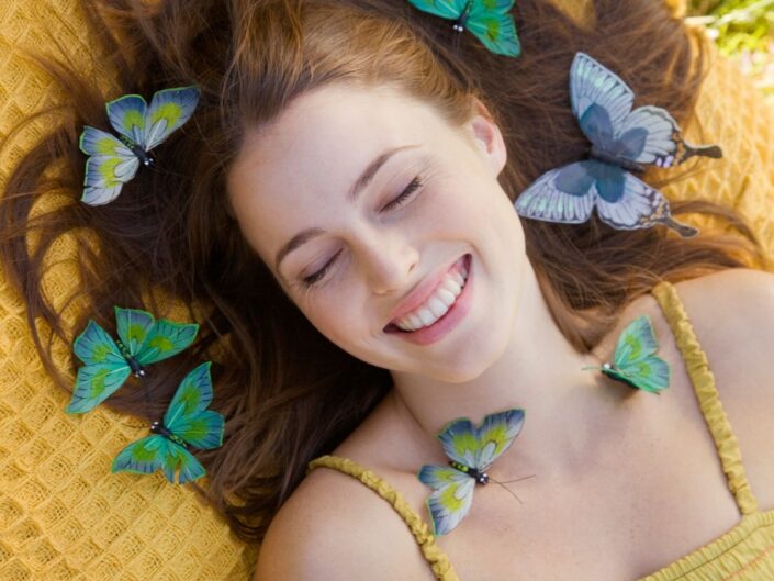 Sárga takarón vörös hajú, mosolygós nő fekszik körülötte kék és zöld pillangók repkednek