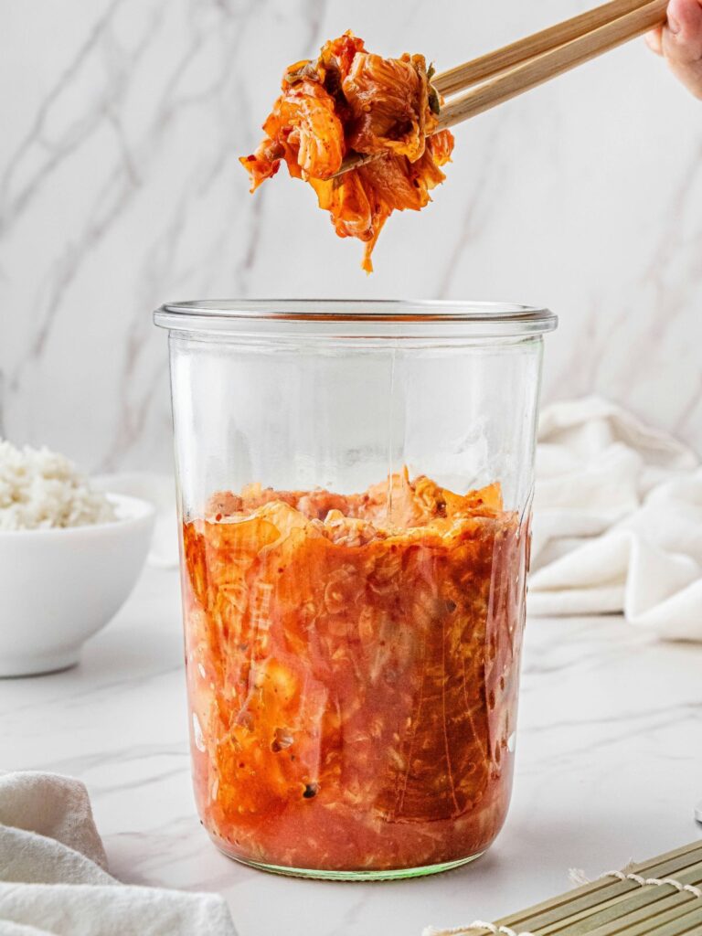 kimchi-mikromuanyag-ellen - Nők Lapja Fehér asztalon kimchi üvegben, evőpálcikával kiemel egy adagot egy kéz