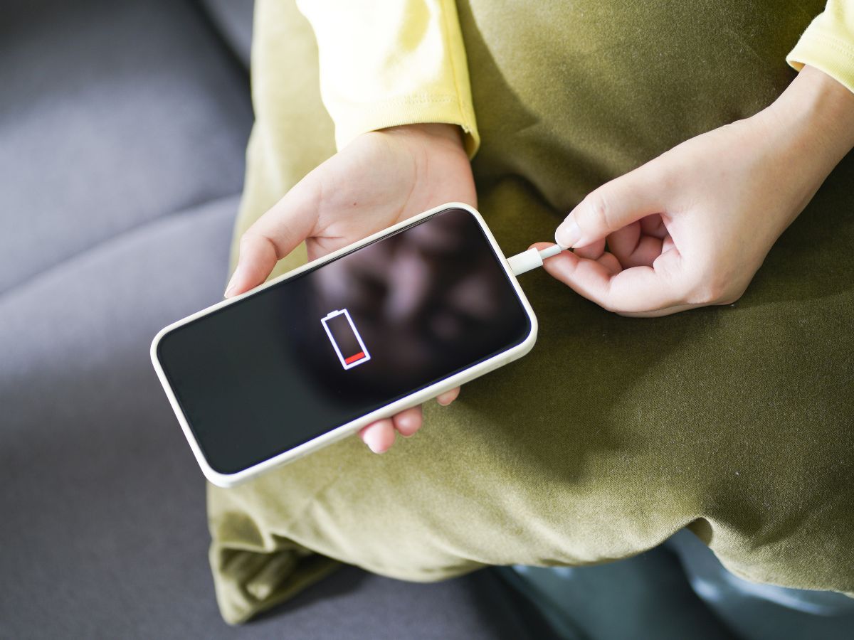 középen egy kézben mobiltelefon, a kijelzőjén lemerült akkumulátor jelzés, jobbról épp töltőre dugják a készüléket, alatta zöld párna