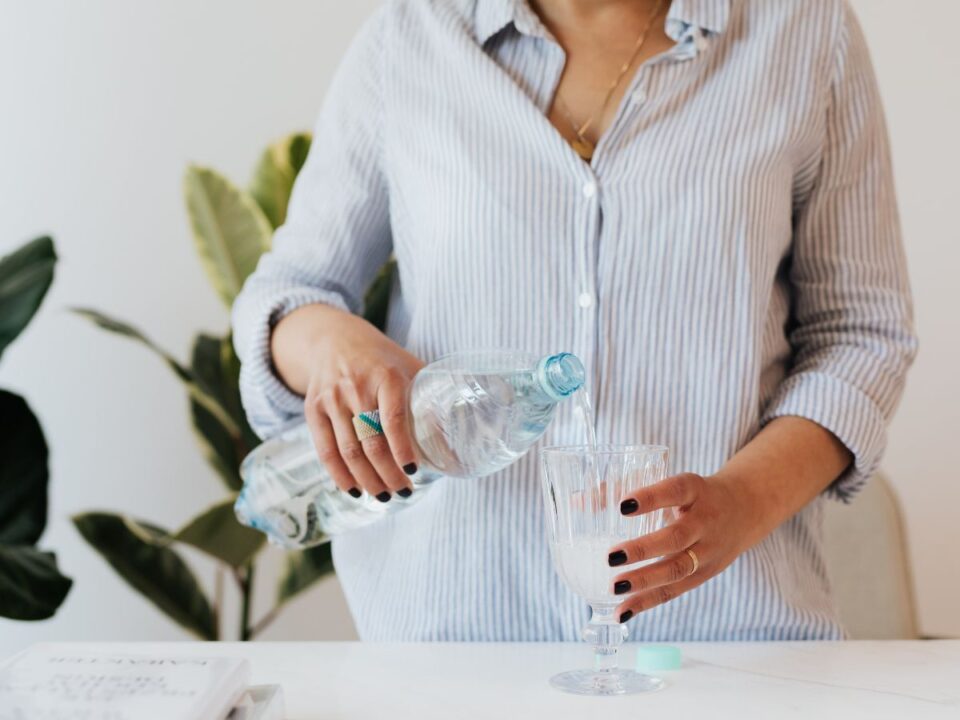 Egy nő világos csíkos ingben műanyag palackból vizet tölt egy pohárba egy asztalnál, háttérben zöld szobanövénnyel, nyugodt, otthoni környezetben.