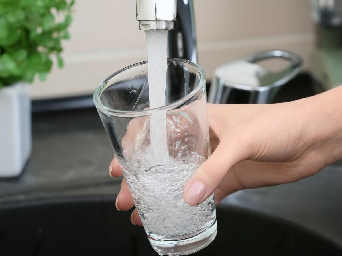 Egy kéz a csap alá tart egy poharat, amelybe friss csapvíz áramlik, buborékokkal telve, konyhai környezetben, háttérben mosogató és vízforraló látható.