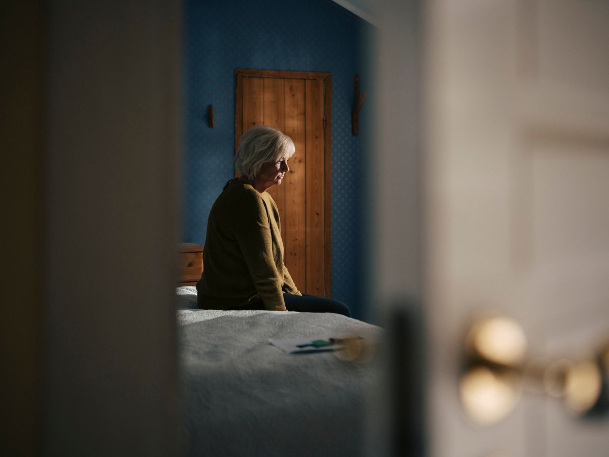 idős nő nyitott ajtó mögött ágy ül mereng