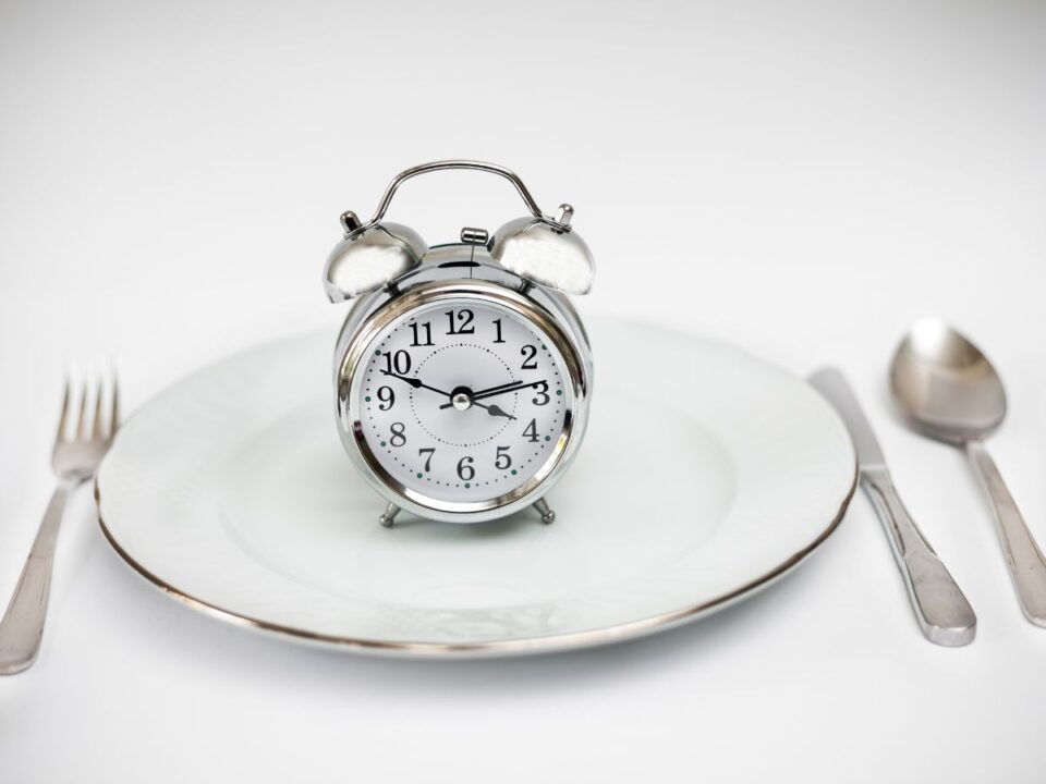 Egy fehér tányér közepén egy klasszikus ébresztőóra áll, mellette villa és kanál. A kép a fémevést szimbolizálja, utalva a szokatlan étkezési szokásokra.