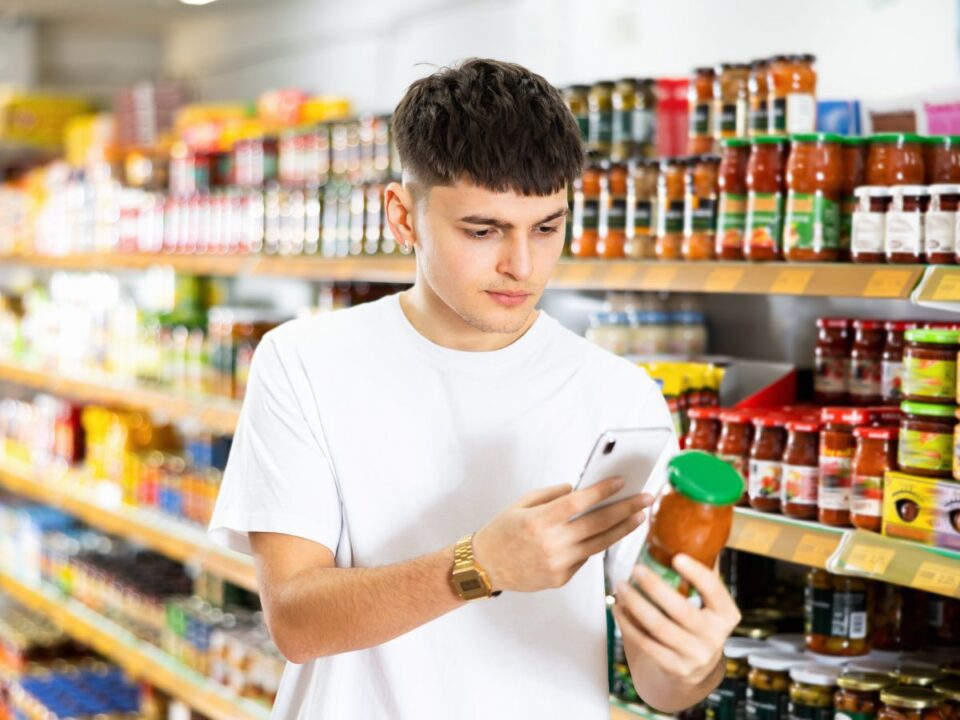 Fehér pólós fiatal férfi, az élelmiszerboltban kézben a mobiltelefonjával egy üveges tésztaszószt azonosít