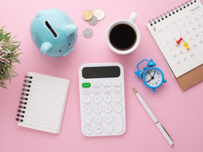 Pasztell háttéren jegyzetfüzet, számológép, kávé, naptár és aprópénz látható, amelyek a tudatos pénzügyi tervezést és a mindennapi kiadások átgondolását jelképezik.
