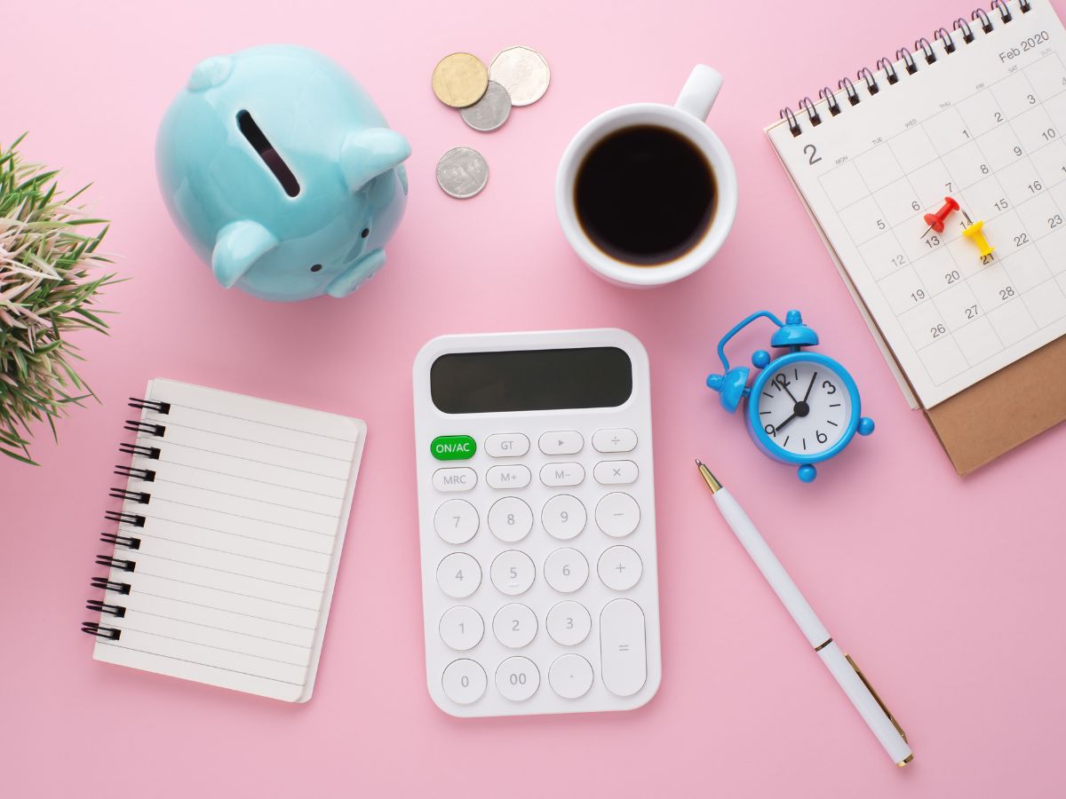 Pasztell háttéren jegyzetfüzet, számológép, kávé, naptár és aprópénz látható, amelyek a tudatos pénzügyi tervezést és a mindennapi kiadások átgondolását jelképezik.