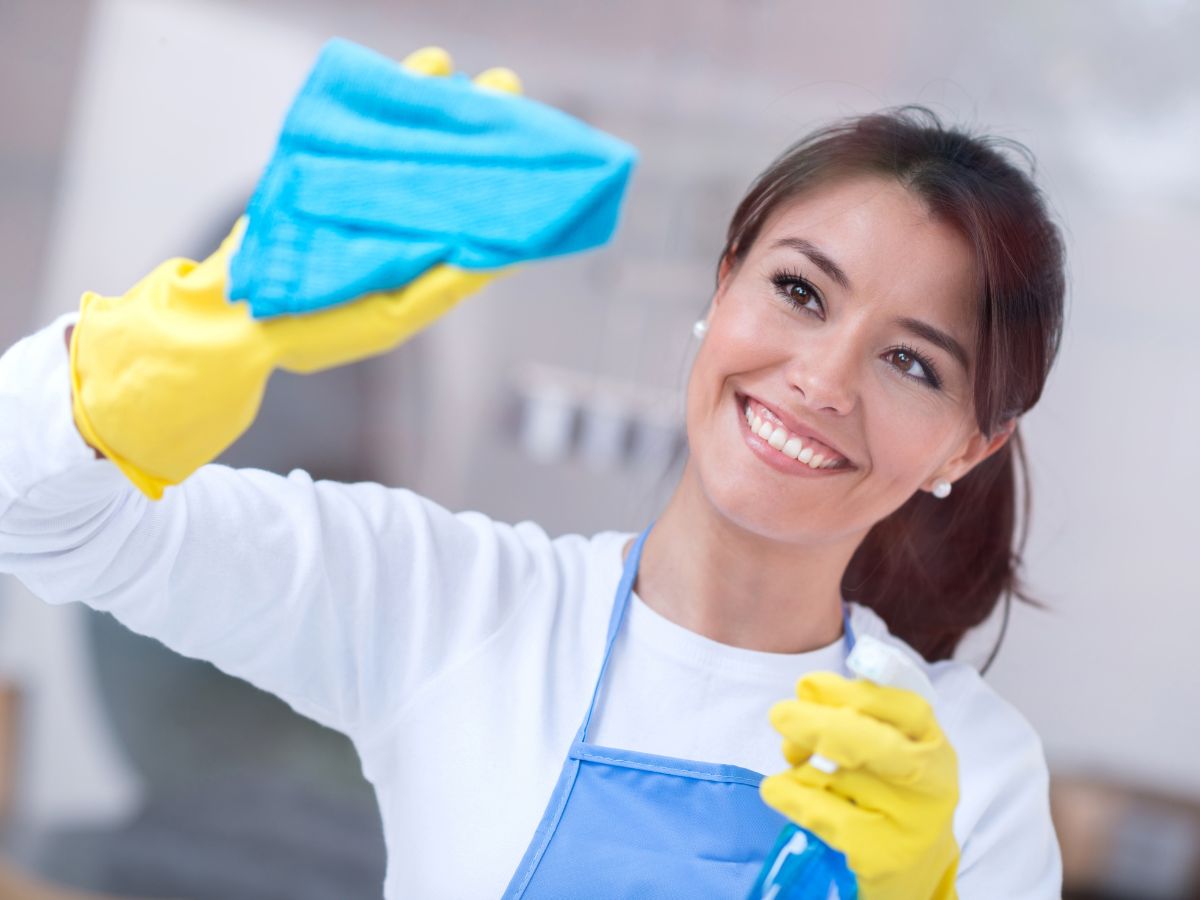 Mosolygó nő sárga gumikesztyűben, kék kendővel töröl egy ablakot; közelről látszik a mozdulata és a tisztítószer, miközben a háttér világos, rendezett otthont sejtet.