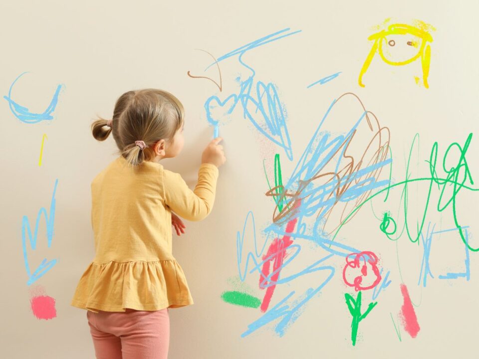 tavaszi-szunet-gyerek-otthon - Nők Lapja Egy kislány színes krétákkal rajzol a falra: firkák, szívek és alakok láthatók, miközben oldalról figyeljük őt, ahogy belemerül az alkotásba.
