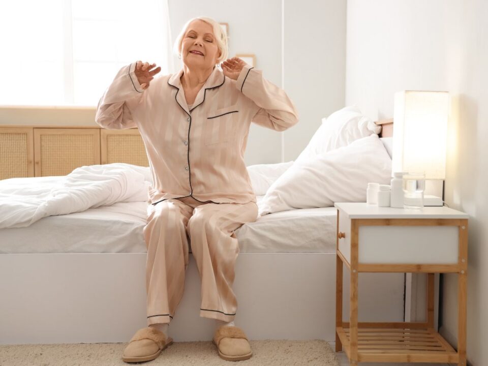 idős nő ágy szélén ülve nyújtózik reggeli fényben