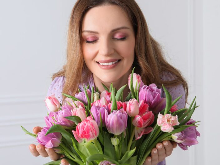 Egy mosolygó nő csukott szemmel tart kezében egy dús, rózsaszín és lila tulipáncsokrot. Az arcát finoman érintik a virágok, a kép hangulata nyugodt és tavaszias.