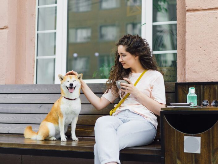 Barna, göndör hajú nő padon ül kezében csészével egy shiba inu mellett, megsimogatja a kutya fejét