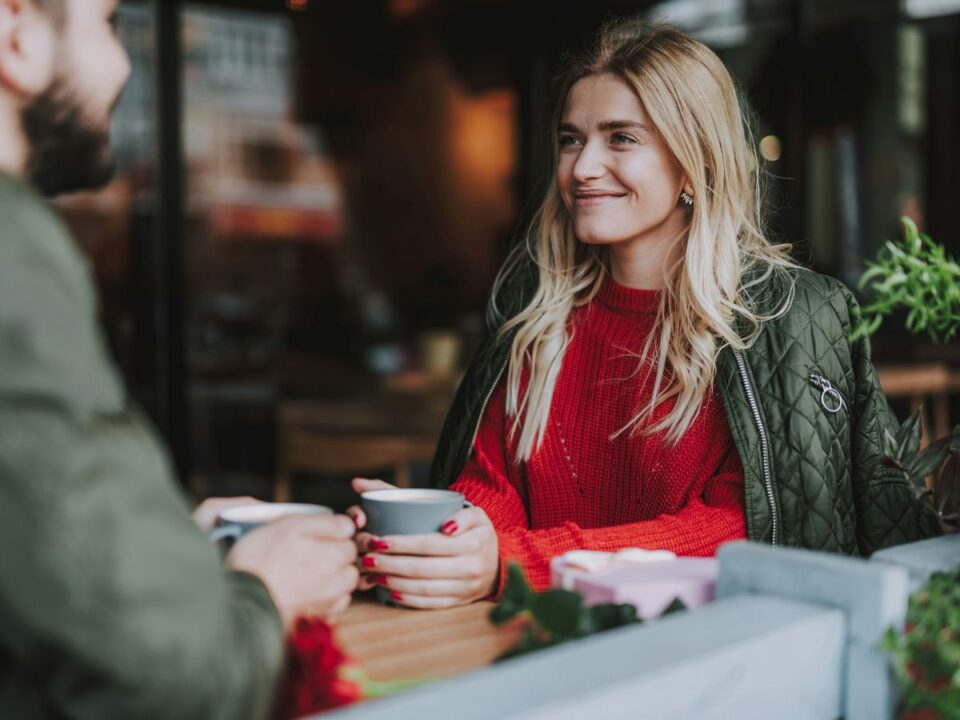 Szőke nő piros felsőben mosolyog csészét fogva a kezében randizás közben