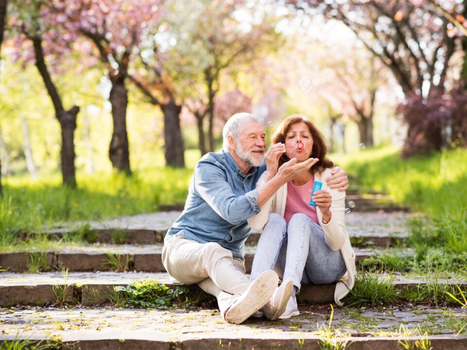 Egy idősebb pár ül a park lépcsőjén, a nő szappanbuborékokat fúj, a férfi mosolyogva nézi; körülöttük zöldellő fák és napsütés.