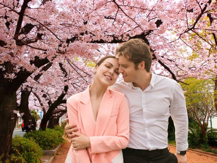 Egy fiatal pár sétál egymást átölelve egy virágzó cseresznyefákkal szegélyezett úton; a nő mosolyogva lehunyja a szemét, a férfi szeretettel néz rá.