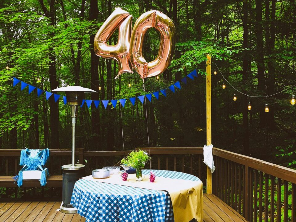 Arany színű 40-es számot formázó lufi születésnapi kék kockás terítővel letakart kerek asztal felett teraszon, háttérben erdő
