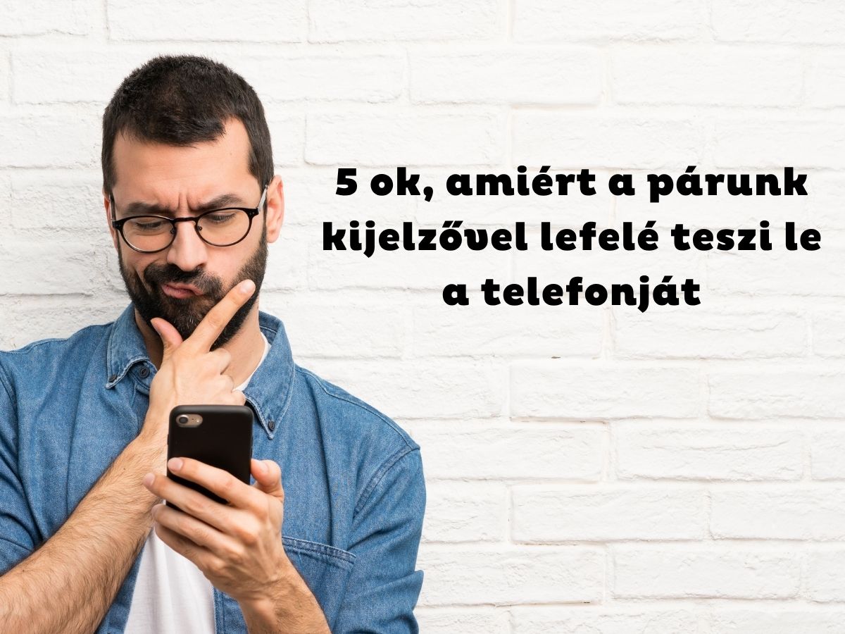 Szemüveges és szakállas férfi farmeringben állát vakarva nézi a telefonját