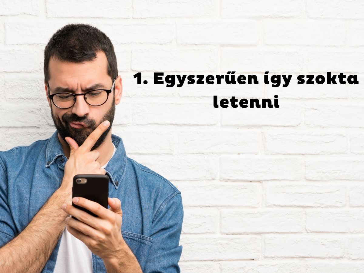 Szemüveges és szakállas férfi farmeringben állát vakarva nézi a telefonját