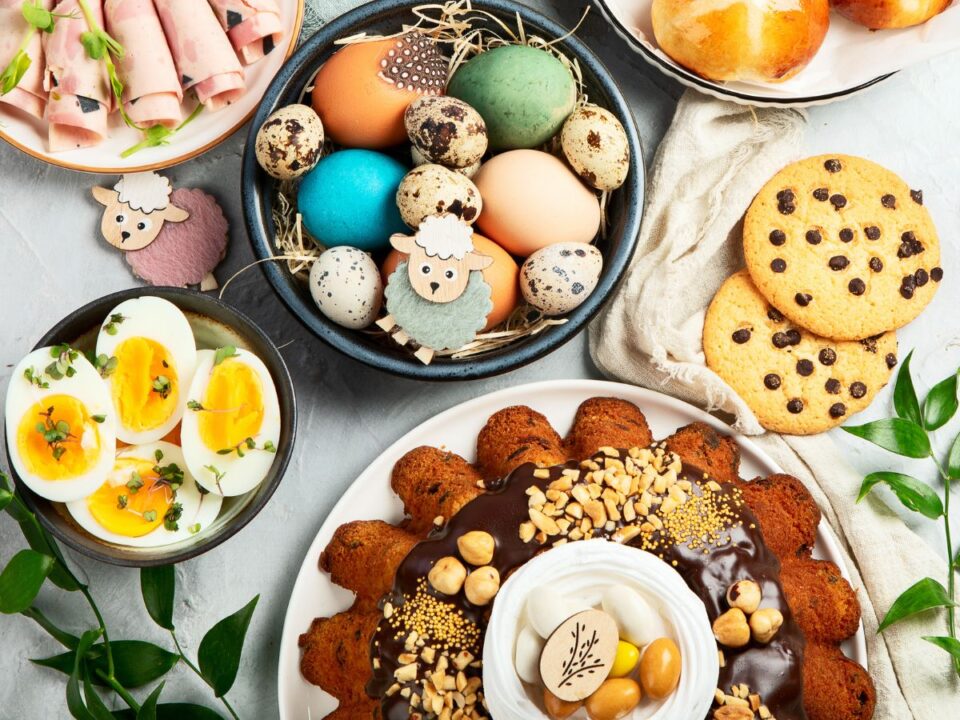 7-penzcsapda-husveti-vendegvaras - Nők Lapja Gazdagon terített húsvéti asztal: festett tojások, sonka, főtt tojás, sütemények és kalácsok színes tálakon, dekorációval és zöld növényekkel.