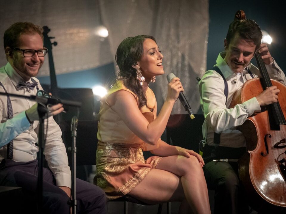 Színpadon ülő Csemer Boglárka Boggie énekesnő mikrofonba énekel, mellette két zenész: egyik csellón játszik, a másik mosolyogva figyel. Meleg fények, elegáns, intim koncert hangulat.