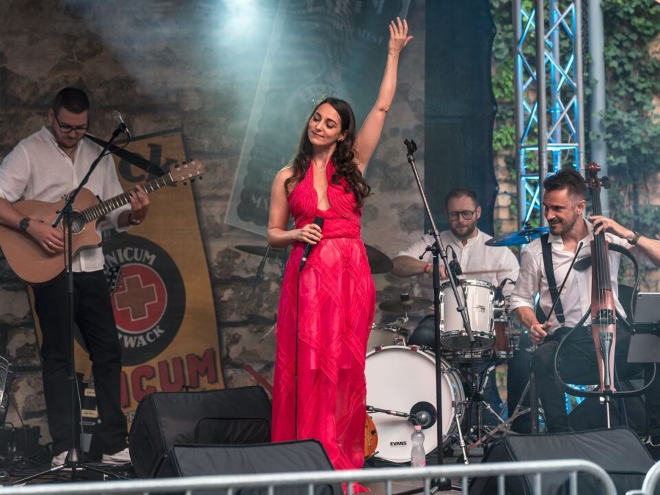 Szabadtéri színpadon piros ruhás énekesnő áll felemelt karral, mögötte zenekar: gitáros, dobos és csellista. Nyári koncert, lendületes, felszabadult hangulat.