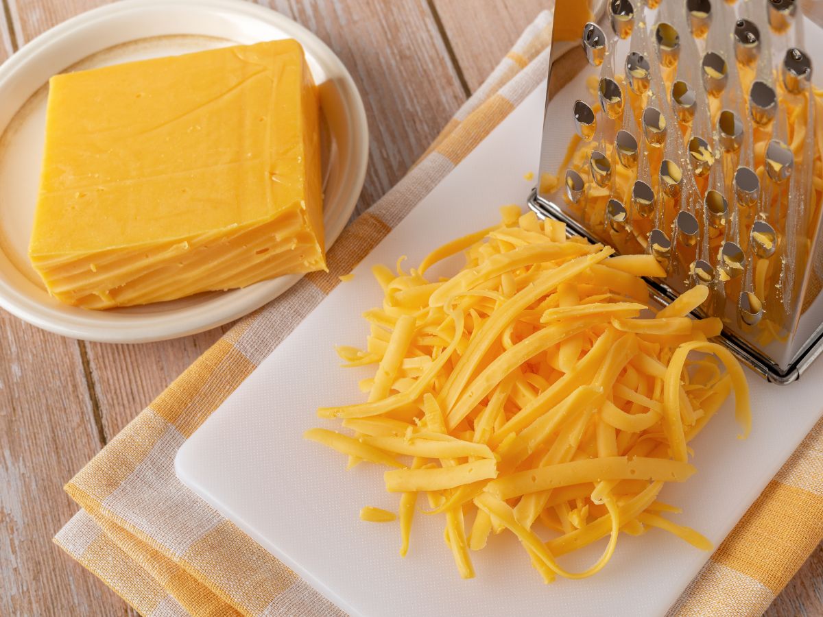 Cheddar sajt tömbben fehér tálban, mellette sajtreszelő és reszelt cheddar fehér deszkán
