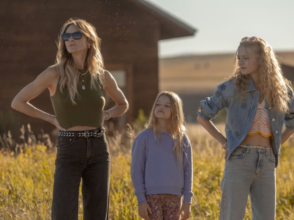 Egy nő két gyerekkel áll egy napsütötte mezőn egy faház előtt. A gyerekek különböző irányba néznek, a nő határozott testtartással figyel.