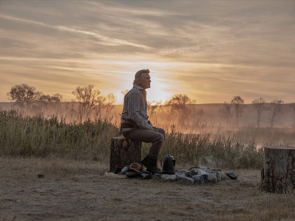 Kurt Russel a The Madison című sorozatban: egy farönkön ül naplementében a mezőn, mellette tűzrakó hely és kávéskanna. A háttérben ködös táj és fák látszanak, a jelenet nyugodt, elmélyült hangulatú.