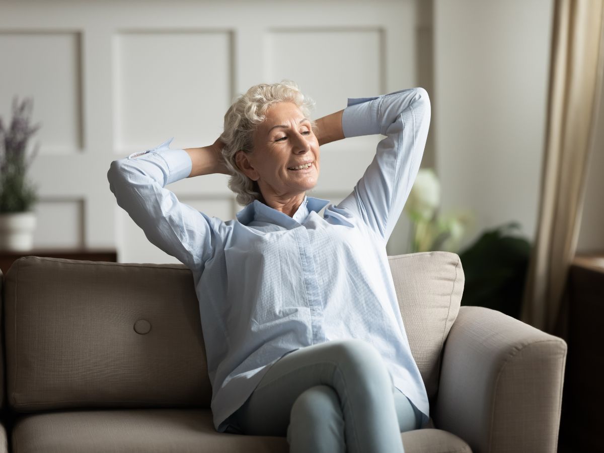 Idősebb, nyugodt nő ül a kanapén, kezeit a tarkóján nyugtatja